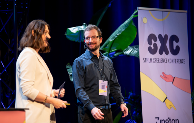 En kvinna och en man står bredvid varandra på en scen med en roll-up i bakgrunden med texten STHLM Xperience Conference